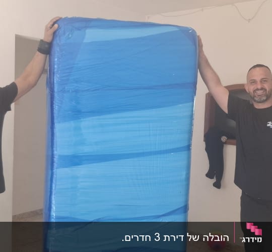 שני אנשים מחזיקים מזרן עטוף בניילון כחול בחדר ריק